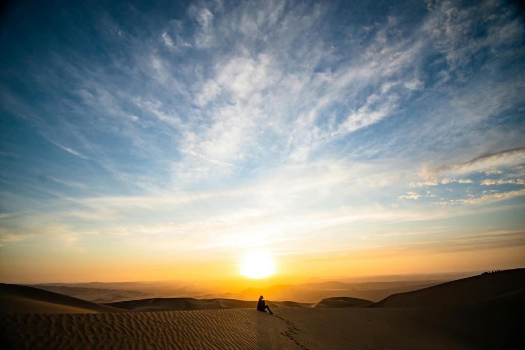 individual sitting atop a sand dune as the sun is going down